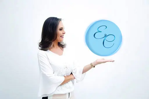 Elizete Caliman de pé, sorrindo, com a mão estendida sob o monograma da sua marca.